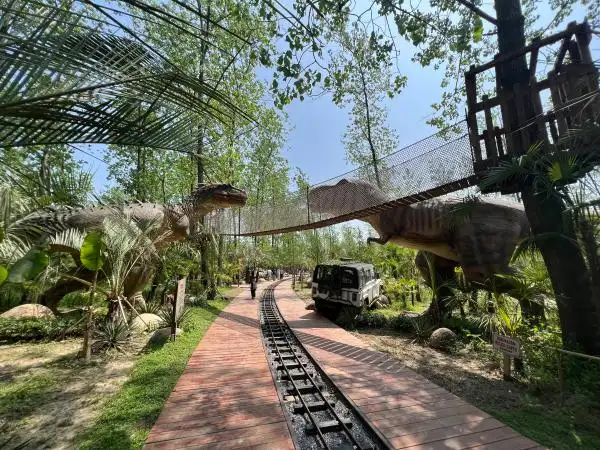 Elevated suspension bridge and railway track at an outdoor dinosaur attraction, featuring large T-Rex animatronics.
